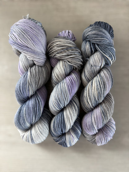 Three skeins of hand-dyed, ethically sourced merino wool yarn in twilight blue, lilac, and muted gray.