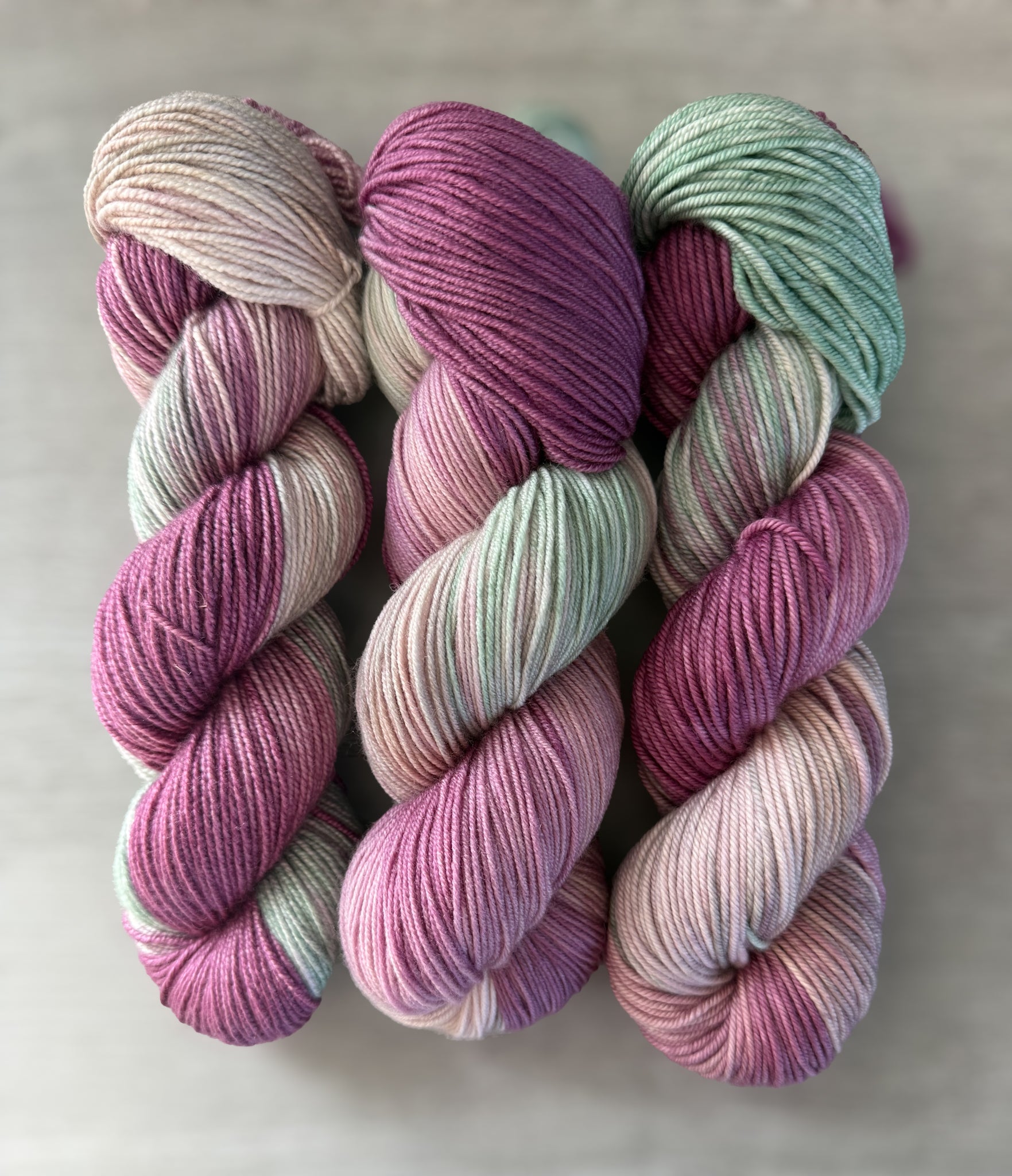 Three skeins of ethically sourced, hand-dyed merino wool yarn in variegated plum, ivory, and sage green.