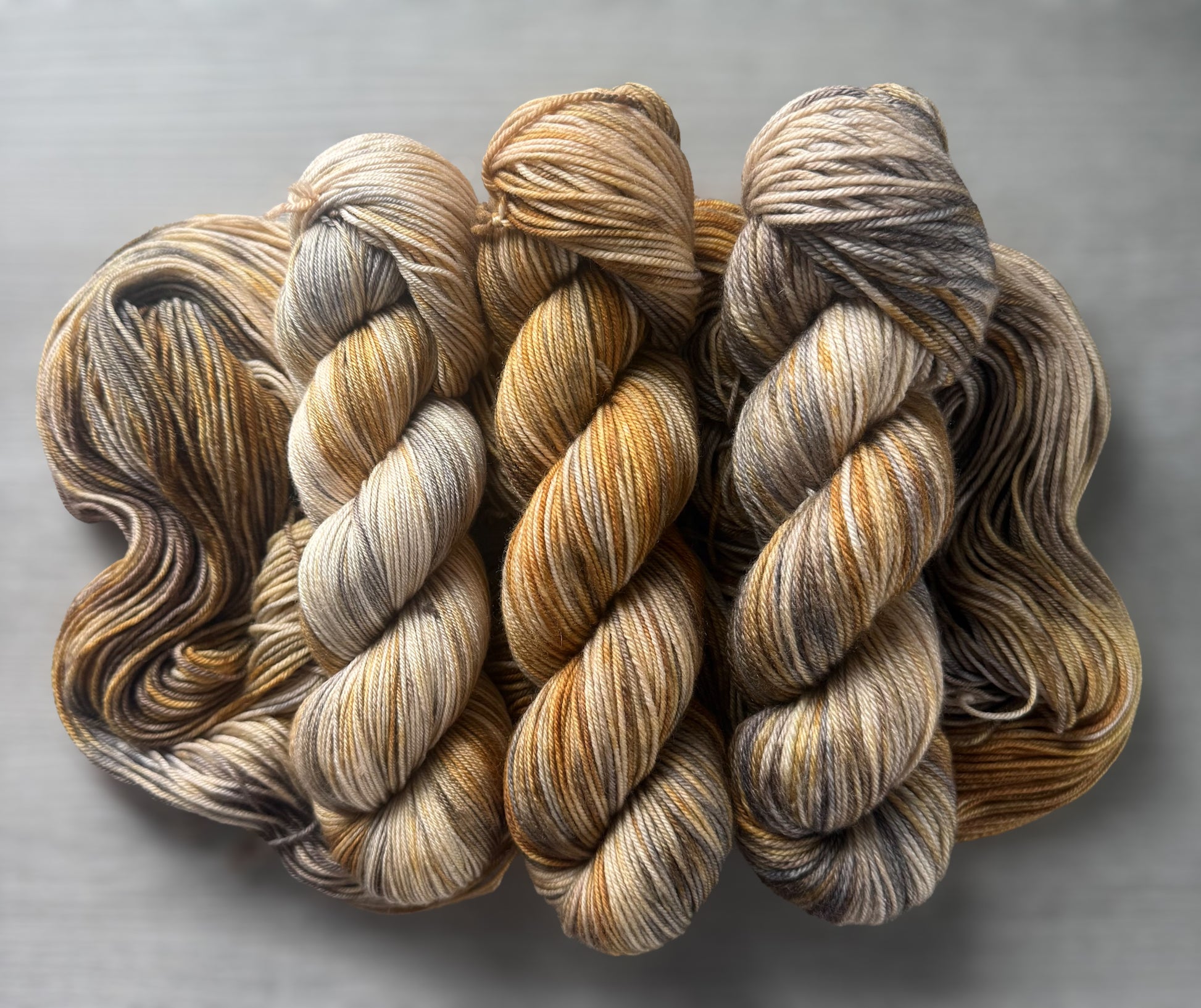 Three skeins of Mesa hand-dyed ethically sourced merino wool yarn with beige, charcoal, taupe, and ocher tones.