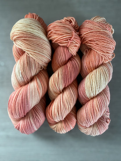 Three skeins of ethically sourced, hand-dyed merino wool yarn in soft peach, ivory, and rose hues.